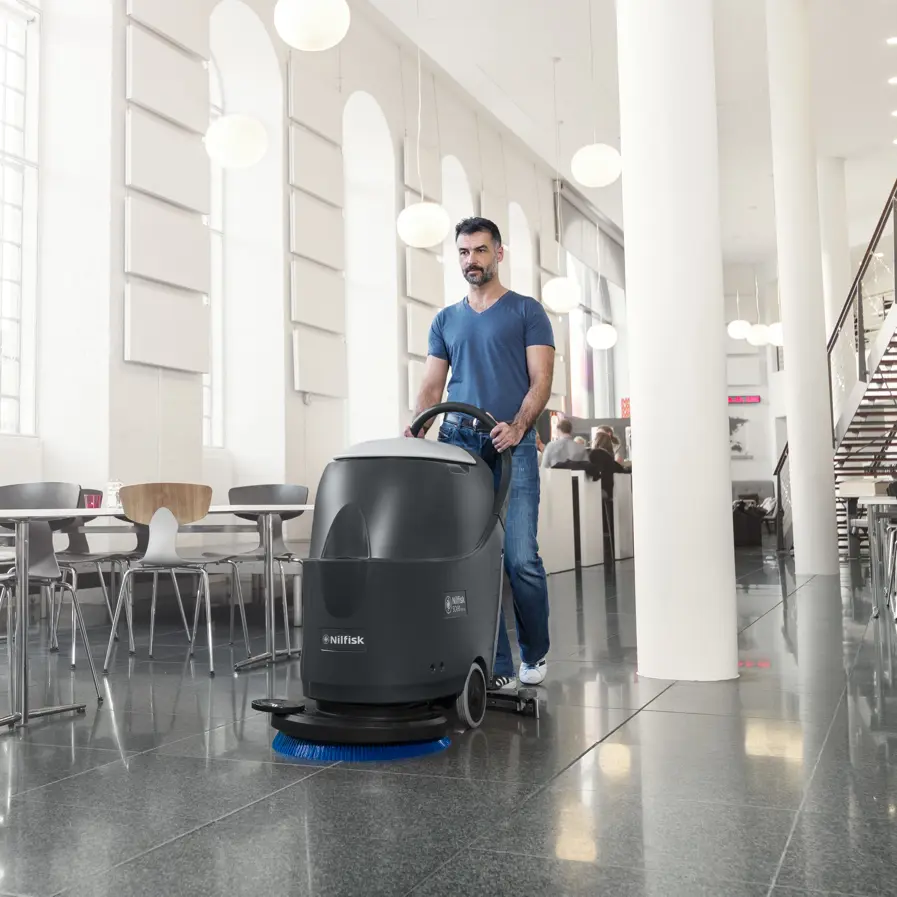 Operator using a Nilfisk walk-behind floor scrubber in a spacious commercial tile floor area.